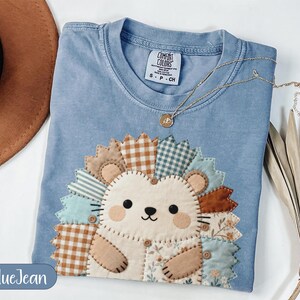 May include: A blue jean colored t-shirt featuring a large, central applique of a hedgehog. The hedgehog is constructed from various fabric patches in shades of brown, cream, and light blue, with visible stitching and small buttons.