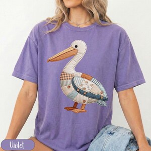 May include: A violet short-sleeved t-shirt featuring a large graphic of a pelican composed of various fabric patches in shades of blue, brown, and white, accented with small brown buttons and visible stitching details across the bird's body.