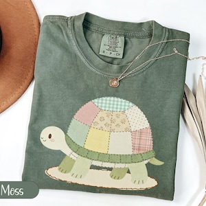 Könnte beinhalten: Ein moosgrünes Kurzarm-T-Shirt mit einer Grafik einer Schildkröte, deren Panzer aus verschiedenen pastellfarbenen Patchwork-Mustern wie Vichy-Karo, Blumen und Schottenkaro besteht. Das T-Shirt hat einen Rundhalsausschnitt und ein Comfort Colors Etikett.