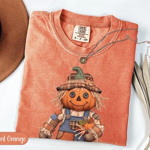 May include: A burnt orange short-sleeve t-shirt featuring a graphic of a scarecrow with a pumpkin head, button eyes, and a plaid hat. The shirt has a Comfort Colors brand label at the neck and a silver-toned chain necklace resting on the fabric.