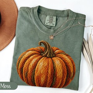 Peut inclure: Un t-shirt à col rond vert mousse avec un grand motif de citrouille texturée orange et marron. Le t-shirt présente une étiquette de marque Comfort Colors à l'intérieur du col, indiquant Comfort Colors, ring spun, 100% cotton, made in Honduras, S P CH.