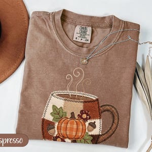 May include: An espresso-coloured t-shirt featuring a central graphic of a coffee mug decorated with a patchwork pattern of pumpkins, acorns, and leaves. The shirt has a crew neckline with a Comfort Colors label and is paired with a silver necklace.