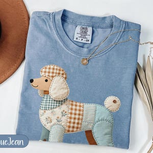 May include: A blue jean colored t-shirt featuring a patchwork poodle graphic. The poodle design is composed of various fabric patterns including brown and white gingham, light blue stripes, and a floral print. The shirt has a crew neckline.