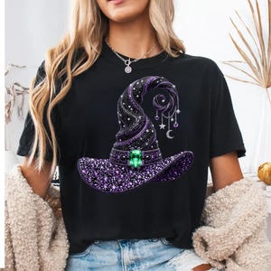 Può includere: Una t-shirt nera con la grafica di un cappello da strega decorato con strass viola e neri, una grande gemma centrale verde smeraldo e ciondoli a forma di stelle e una mezzaluna appesi alla punta del cappello.