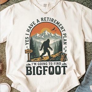 May include: An off-white t-shirt featuring a graphic of a silhouette of Bigfoot walking in a forest with mountains in the background. The text reads: YES I HAVE A RETIREMENT PLAN I'M GOING TO FIND BIGFOOT. A wooden sign and a backpack are visible.