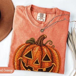 Peut inclure: Un t-shirt à manches courtes orange brûlé avec un grand motif de citrouille d'Halloween à l'effet texturé brodé. Le t-shirt présente un col rond avec une étiquette de marque Comfort Colors indiquant la taille S-CH. Un collier argenté est posé.
