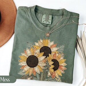 Może przedstawiać: Koszulka z krótkim rękawem w kolorze mchu z dużą, pikowaną aplikacją słonecznika z przodu. T-shirt ma okrągły dekolt z metką Comfort Colors. Na tkaninie spoczywa delikatny złoty naszyjnik z małym, okrągłym wisiorkiem.