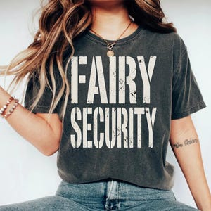 May include: A charcoal grey short-sleeve t-shirt featuring the text FAIRY SECURITY printed in a distressed, off-white, sans-serif font across the chest. The garment has a crew neckline and a relaxed fit, showcasing a weathered, vintage-inspired texture.