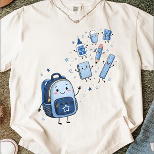 Op de afbeelding: Een off-white t-shirt met korte mouwen en een grafische print van een lachende blauwe rugzak omringd door geanimeerde schoolspullen, waaronder een lijmflesje met het label GLUE, een notitieboekje, een potlood, een gum en een liniaal.
