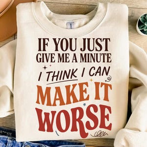 Könnte beinhalten: Ein cremefarbenes Sweatshirt mit Rundhalsausschnitt und dem dunkelbraunen, orangen und roten Text: IF YOU JUST GIVE ME A MINUTE I THINK I CAN MAKE IT WORSE. Das Design enthält kleine dekorative Stern- und Wirbelgrafiken auf der Vorderseite.