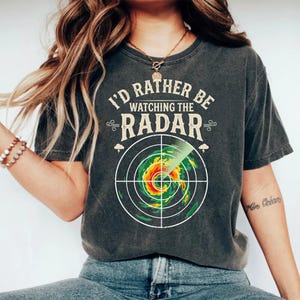 Könnte beinhalten: Ein anthrazitfarbenes Kurzarm-T-Shirt mit dem Text I'D RATHER BE WATCHING THE RADAR in cremefarbener Schrift über einer Grafik eines Wetterradarschirms, der ein wirbelndes Sturmsystem in Grün-, Gelb- und Rottönen anzeigt.