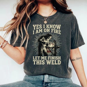 Peut inclure: Un t-shirt gris anthracite à manches courtes avec un motif représentant un soudeur portant un masque de protection travaillant sur un tuyau métallique. Le texte indique YES I KNOW I AM ON FIRE LET ME FINISH THIS WELD, avec un motif en rayons de soleil.