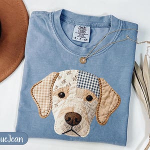 May include: A blue crew neck t-shirt featuring a patchwork dog face applique made of various beige, white, and blue patterned fabrics. The shirt has a Comfort Colors label at the neck and a gold-toned pendant necklace resting on the fabric.