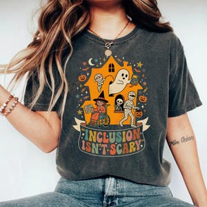 Puede incluir: Una camiseta gris carbón con un gráfico colorido de Halloween que muestra una casa embrujada con un fantasma, una momia, un esqueleto y un niño en silla de ruedas. El texto "INCLUSION ISN'T SCARY" aparece en una tipografía retro en una banda.