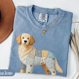 May include: A blue crew neck t-shirt featuring a graphic of a golden retriever dog composed of various fabric patches in plaid, floral, and solid patterns. The shirt has a Comfort Colors brand label at the neck and is size S-CH.