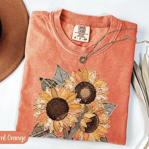 Könnte beinhalten: Ein kurzärmeliges T-Shirt in gebranntem Orange mit einem großen Applikationsdesign aus drei Sonnenblumen mit strukturierten Stoffblütenblättern in Gelb-, Braun- und Grüntönen. Das T-Shirt hat einen Rundhalsausschnitt mit einem Comfort Colors Etikett.