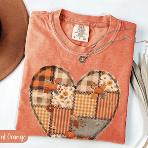 May include: A burnt orange t-shirt featuring a large heart-shaped graphic composed of various patchwork patterns including plaid, stripes, and floral prints. The design includes small orange pumpkins and autumn leaves with visible black zigzag stitching.