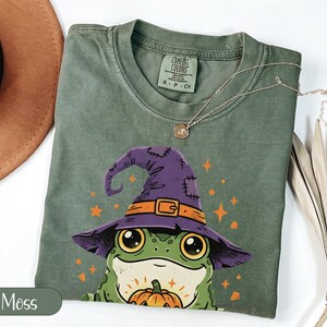 May include: A moss green short-sleeve t-shirt featuring a graphic of a green frog wearing a purple witch hat and holding a small orange pumpkin. The shirt has a crew neckline and a Comfort Colors brand label printed on the inside collar area.