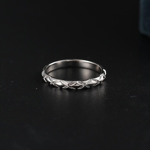 May include: A silver ring with a repeating leaf-like pattern around the band. The ring is made of a shiny, reflective metal and sits on a dark surface. The ring's design is symmetrical and detailed, showcasing a delicate floral motif.