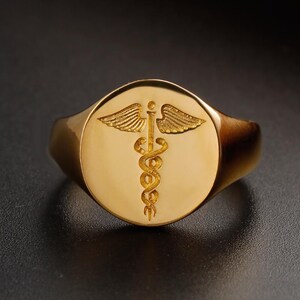 May include: A gold signet ring featuring the Rod of Asclepius symbol, a serpent-entwined staff with wings. The ring has a polished finish and a rounded face. The ring is a medical or apothecary themed item.
