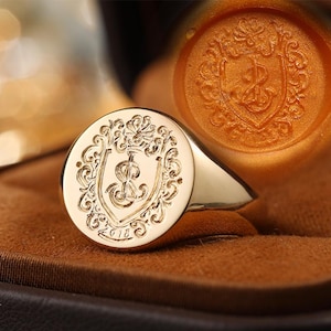 May include: A gold signet ring with a detailed crest design. The ring features a round face with an ornate shield, dollar sign, and the year "2018" engraved. The ring is displayed on a brown surface.