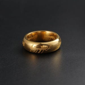 May include: A gold-colored ring with engraved text. The ring has a smooth, polished surface and a classic band design. The engraved text appears to be in a stylized script. The ring is set against a dark background.