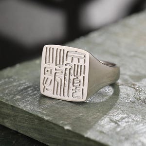 May include: A silver signet ring with a square face featuring an engraved design. The ring is made of a shiny, metallic material and rests on a textured, gray surface. The engraved design appears to be an intricate pattern of lines and shapes.