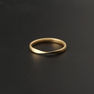 May include: A gold-colored ring with a twisted design. The ring is set against a dark background, highlighting its smooth, matte finish. The band has a subtle curve, adding a unique touch to the jewelry piece.