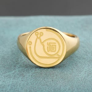 May include: A gold-colored signet ring with a circular face. The face features a detailed engraving of a snail within a circular border. The ring has a polished finish and a classic, rounded band.
