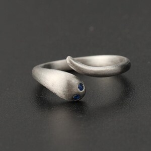 Può includere: Un anello color argento a forma di serpente, con finitura in metallo spazzolato. La testa del serpente presenta due piccole gemme blu rotonde come occhi. L'anello si curva, creando un design unico ed elegante.