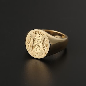 May include: A gold signet ring with an oval face featuring a detailed crest design. The crest includes a crown, shield, and two flanking lion figures. The ring has a polished, smooth band and a bright, reflective finish.