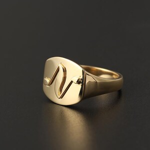 Puede incluir: Un anillo de sello dorado con una cara cuadrada. La cara presenta un diseño curvo estilizado. El anillo es de metal pulido y brillante, y está sobre una superficie oscura. Es una pieza de joyería.