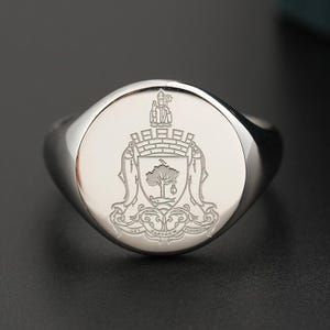 Puede incluir: Anillo de sello plateado pulido con un escudo grabado detallado. El escudo presenta un árbol, un pez y una figura sobre un castillo, con las palabras "LET GLASGOW FLOURISH" en la base. El anillo tiene una cara ovalada lisa.