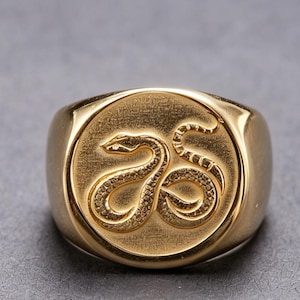May include: A gold-toned signet ring featuring a circular face with an embossed, detailed snake design. The snake is coiled in a curved shape, displaying textured scales across its body. The ring has a polished, smooth finish on the band.