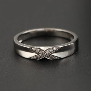 Può includere: Un anello d'argento con un design a forma di X. L'anello presenta una fascia lucida e un dettaglio centrale a forma di X ornato da piccole pietre preziose. L'anello è presentato su uno sfondo scuro.