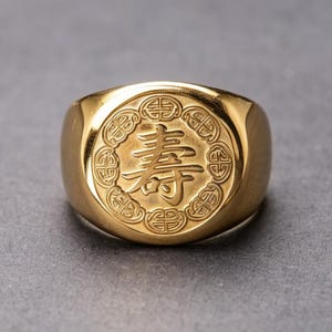 Op de afbeelding: Een goudkleurige zegelring met een cirkelvormig voorvlak met in het midden een gegraveerd Chinees karakter voor een lang leven, omringd door een decoratieve rand van herhalende geometrische knooppatronen op een gepolijst metalen oppervlak.