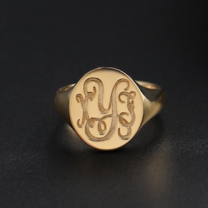 May include: A gold signet ring with an engraved monogram. The ring has a polished finish and a rounded face with the initials in a flowing script. The ring is set against a dark background.