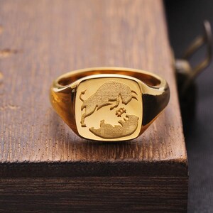 以下が含まれることがあります： 金色のリングで、四角い面に雄牛と熊のデザインが施されています。 リングは研磨仕上げで、木製の表面に置かれています。 雄牛は熊の上にあり、その間にシンボルがあります。 シグネットリングスタイルです。