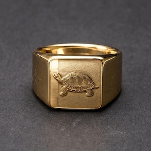 May include: A gold-toned signet ring featuring a flat, rectangular face with a detailed, engraved turtle design. The ring has a polished, reflective finish on the band and a textured, matte-like surface on the central square panel.
