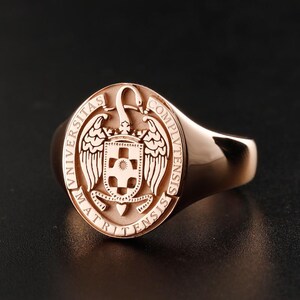 Op de afbeelding: Rosegouden zegelring met een gedetailleerd universitair wapen. Het wapen toont een schild, vleugels en de woorden "UNIVERSITAS COMPLUTENSIS MATRITENSIS" langs de rand. De ring heeft een gepolijste afwerking en een klassiek ontwerp.