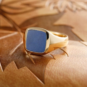 May include: A gold ring with a square, blue stone set in a gold band. The ring has a polished finish and is displayed on a textured, brown surface. The ring is a piece of jewelry.