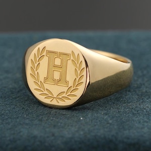 May include: A gold signet ring with a polished finish. The ring features a raised letter "H" within a laurel wreath design on the face. The ring is set against a dark teal background.