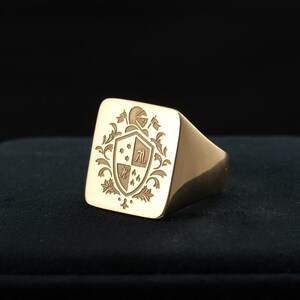 Könnte beinhalten: Goldener Siegelring mit quadratischer Oberfläche. Die Oberfläche ist mit einem Wappen mit Schild, Helm und floralen Elementen graviert. Der Ring hat eine polierte Oberfläche und ein dickes Band.