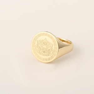 Może przedstawiać: Złoty sygnet z okrągłą tarczą z grawerowanym wzorem mitycznego stworzenia. Pierścionek ma polerowane wykończenie i grubą obręcz. Pierścionek to klasyczny element biżuterii.