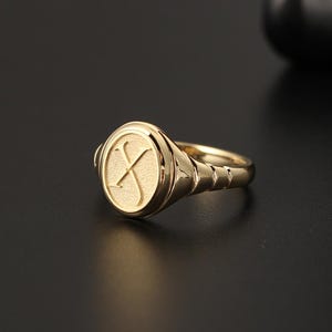 May include: A gold signet ring with an oval face featuring the letter "X". The ring has a polished finish and a simple band with ribbed detailing. The ring is set against a dark background.