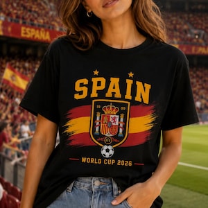 May include: A black short-sleeve t-shirt featuring the word SPAIN in yellow letters above a graphic of the Spanish coat of arms, a red and yellow brushstroke flag, a soccer ball, and the text WORLD CUP 2026 printed in yellow and red.