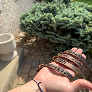 May include: A stack of brown leather bracelets with engraved text. The bracelets are held in a hand, with the text including phrases like "Hinini" and "Here am I. Send me". The bracelets have a rustic, worn appearance.