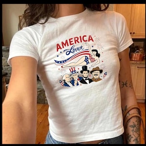 Könnte beinhalten: Weißes T-Shirt mit dem Text "AMERICA Lover" in Rot und Blau, mit einer Grafik einer Frau in patriotischer Kleidung und Illustrationen historischer Persönlichkeiten. Das Design enthält Sterne und Feuerwerk.