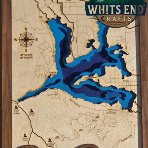 May include: Wooden map of Pineview Reservoir in Utah, featuring a detailed topographical design with varying shades of blue to represent water depth. The map includes roads, landmarks, and the text "WHITS END CRAFTS". The frame is made of dark wood.