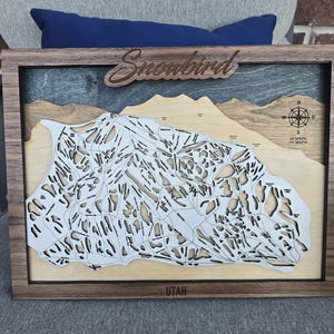 May include: Wooden framed map of Snowbird, Utah, with a detailed ski run layout. The map is laser-cut with a light-colored wood, set against a blue and tan background. The word "Snowbird" is carved at the top.